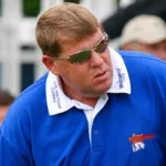 John Daly wearing a blue polo shirt and sunglasses at a golf event, featured in coverage of John Daly Net Worth and his golf career.