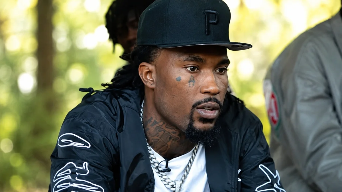 Hunxho Net Worth portrait showing the rapper wearing a black cap, jacket, and silver chains outdoors