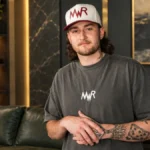 Austin Snell Net Worth portrait showing Austin Snell in a dark T-shirt and white cap in a modern lounge.