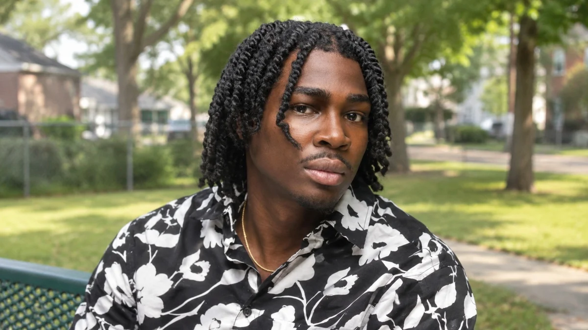 Kam Patterson Net Worth early life image showing Kam Patterson wearing a black-and-white floral shirt while sitting in a park.